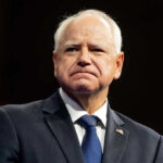 Walz Pulls Race Card in Pathetic Fraud Scandal Defense Man in suit with a U.S. flag pin.