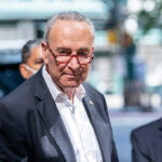 Schumer’s BRUTAL Body Camera Flip-Flop Exposed Man in suit and red glasses standing outside.