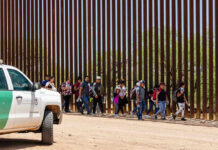 Forced To Deport Despite Living in U.S Since 4 Months Old People walking beside tall fence and border patrol vehicle.