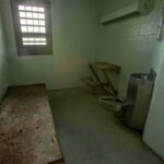 Empty jail cell with bed, barred window, and stainless steel toilet
