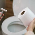 Hands holding a bidet sprayer and toilet paper near an open toilet