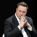 Elon Musk seated in a dark setting with hands steepled near his face