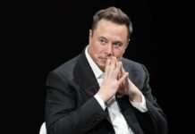 Elon Musk seated in a dark setting with hands steepled near his face