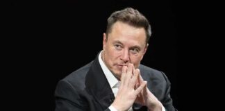 Elon Musk seated in a dark setting with hands steepled near his face