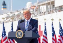Trump Slashes Biden Rule – Says He Can Do It Better Donald Trump speaking at a podium with American flags and a ship in the background