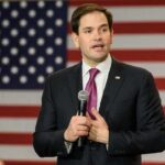 man in suit speaking with microphone before American flag