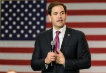 man in suit speaking with microphone before American flag