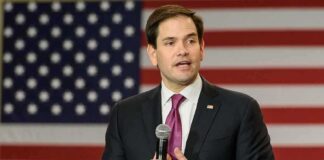 man in suit speaking with microphone before American flag