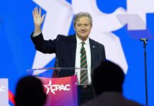 John Neely Kennedy waves while speaking at a podium on a brightly lit stage