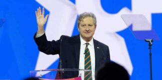 John Neely Kennedy waves while speaking at a podium on a brightly lit stage